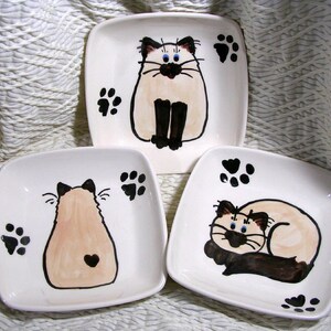 May include: Three white ceramic square plates with hand-painted Siamese cat designs. The first plate features a cat sitting with its tail curled around its legs. The second plate features a cat sitting with its back to the viewer. The third plate features a cat lying down with its head resting on its paws.