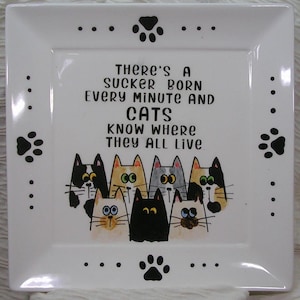 May include: A square white ceramic plate featuring black paw prints along the edges and a central illustration of seven colorful cats. The text reads: THERE'S A SUCKER BORN EVERY MINUTE AND CATS KNOW WHERE THEY ALL LIVE. The cats have varied markings.