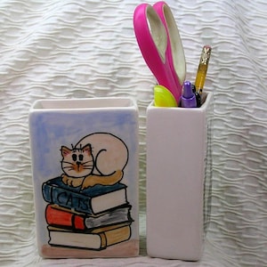 May include: Two ceramic pen holders. One is rectangular with a light blue background and a cat illustration on a stack of books. The books are labeled "CATS". The other holder is white and contains pink scissors, a pencil, and a purple pen.