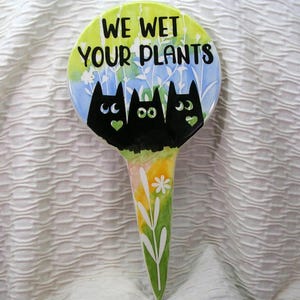 May include: A ceramic garden stake with the text "WE WET YOUR PLANTS". The stake features three black cat silhouettes with green heart accents, set against a backdrop of a green and blue watercolor design. The stake has a long, pointed end with floral details.