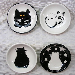 May include: Four small, round ceramic dishes with black cat designs. Each dish features a different cat illustration: a black cat with yellow eyes, a sleeping cat, a cat's silhouette, and a cat silhouette with stars.