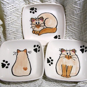 May include: Three white ceramic square dishes with hand-painted images of Siamese cats. The cats are in different poses, one is curled up, one is sitting, and one is facing away from the viewer. Each dish has black paw prints painted on the edge.