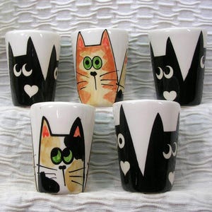 May include: A collection of five ceramic shot glasses. Each glass features a different cat design, with black and white, orange, and calico cat faces. The glasses have a white background and are decorated with hearts and other playful details.