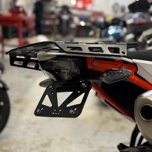 May include: Black metal motorcycle tail tidy with a license plate holder, turn signals, and a luggage rack. The tail tidy is mounted on an orange and white motorcycle.