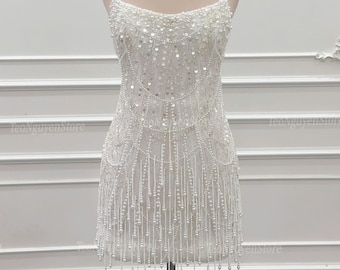 May include: A white, beaded mini dress with a strapless design, displayed on a mannequin. The dress features intricate beadwork and dangling strands of beads. A gold crown sits atop the mannequin's head, adding a regal touch.