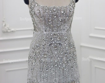 Könnte beinhalten: Ein silbernes, perlenbesetztes Minikleid mit quadratischem Ausschnitt und Fransen. Das Kleid ist stark mit glitzernden Kristallen und Perlen verziert und schafft ein glamouröses Design. Das Kleid wird auf einer Schaufenquin präsentiert.