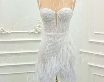 Könnte beinhalten: Ein weißes Minikleid mit einem bestickten Oberteil und einem Federrock. Das Kleid hat Spaghettiträger und einen tiefen Ausschnitt. Auf dem Kopf der Schaufensterpuppe sitzt eine goldene Krone. Das Kleid wird auf einem goldenen Ständer präsentiert.