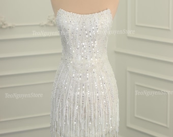 Könnte beinhalten: Ein weißes, trägerloses Minikleid mit einem perlenbesetzten Oberteil und Fransen. Das Kleid ist aus einem durchsichtigen Stoff gefertigt und hat eine figurbetonte Silhouette.