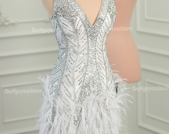 Könnte beinhalten: Ein weißes, ärmelloses Minikleid mit tiefem V-Ausschnitt. Das Kleid ist mit silbernen Pailletten und Perlen verziert und hat einen Saum mit Federn.