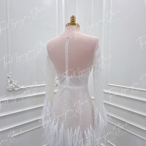May include: A white mini dress with long sleeves and a feather trim. The dress is made of a sparkly fabric and has a fitted silhouette.