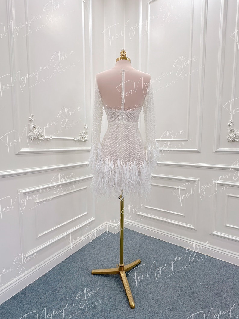 May include: A white mini dress with long sleeves and a feather trim. The dress is beaded and has a fitted silhouette.