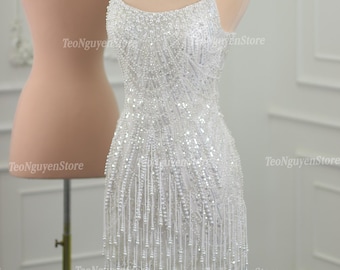 May include: A white strapless mini dress with a beaded bodice and fringe detail. The dress is made of a sheer fabric and has a fitted silhouette.