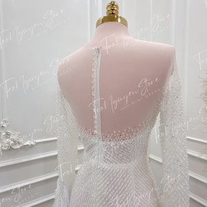May include: A white beaded long-sleeved dress with a feather trim. The dress has a fitted bodice and a flowing skirt. The dress is on a mannequin.