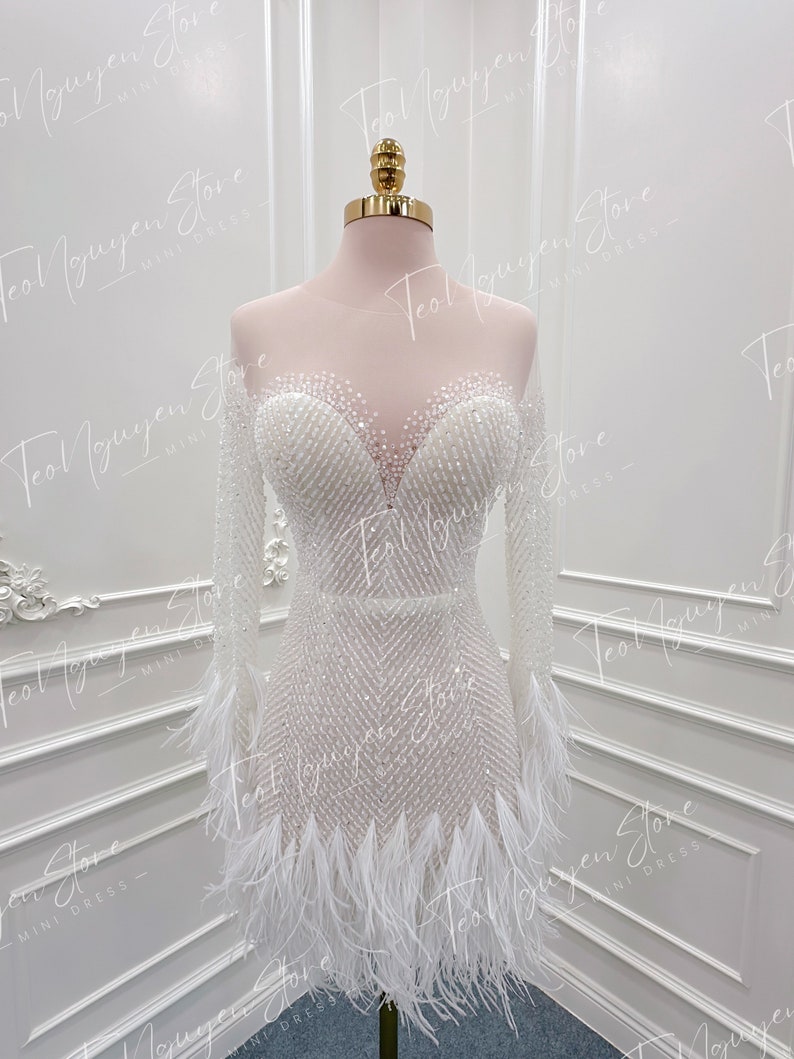 May include: A white mini dress with long sleeves and a feather trim. The dress is covered in small white beads and has a sweetheart neckline.
