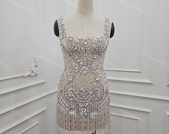 Könnte beinhalten: Ein beiges, verziertes Minikleid mit quadratischem Ausschnitt und Trägern. Das Kleid ist stark mit glitzernden Kristallen und Perlen verziert und hat einen Fransenbesatz. Das Kleid wird auf einer Schaufensterpuppe präsentiert.