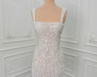 May include: A white, beaded mini dress with a square neckline and spaghetti straps. The dress is adorned with sequins and dangling beaded fringe. A gold crown sits atop the mannequin's head. The dress is displayed on a wooden dress form.