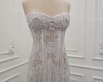 May include: A strapless white gown with intricate beadwork and lace detailing. The bodice features a sweetheart neckline adorned with crystals and delicate beaded strands. A gold crown sits atop the mannequin's head.