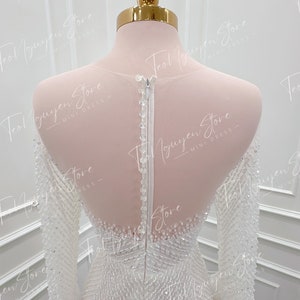 May include: A white beaded long-sleeved dress with a zipper closure in the back. The dress has a fitted silhouette and features feather trim on the sleeves.