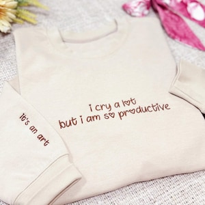 Może przedstawiać: Beżowa bluza z haftem z tekstem "I cry a lot but I am so productive" i "It's an art" na przodzie i rękawie.