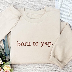 May include: A beige sweatshirt with the text "born to yap." embroidered in brown on the front.