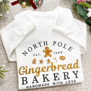 May include: White long-sleeve sweatshirt with a gold embroidered design that says "North Pole Est. 1909 Gingerbread Bakery Handmade with Love". The design includes gingerbread men, candy canes, and stars.