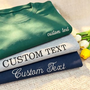 Op de afbeelding: Drie opgevouwen sweatshirts in groen, grijs en marineblauw. Elk sweatshirt heeft de woorden "custom text" of "CUSTOM TEXT" erop geborduurd. De sweatshirts zijn op elkaar gestapeld, met een paar bloemen op de achtergrond.