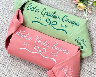 May include: Two folded sweatshirts with embroidered sorority names and bows. The top sweatshirt is green with the text "Beta Epsilon Omega EST. 2025" and the name "Brittany". The bottom sweatshirt is pink with the text "Alpha Theta Sigma EST. 2025" and the name "Ellison".