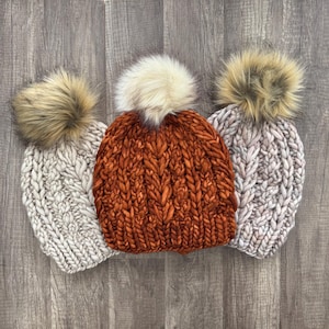 May include: Three chunky knit beanies with faux fur pom-poms. The beanies are in shades of cream, rust orange, and light grey. The hats are arranged on a wooden surface, showcasing their textured knit design and fluffy pom-poms.