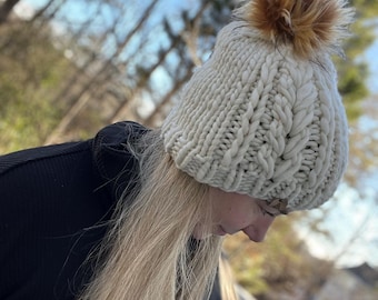 May include: A chunky knit beanie in an off-white color featuring a thick cable knit pattern and a fluffy, multi-toned brown faux fur pom-pom attached to the top. The hat is constructed from thick yarn with a ribbed brim and a textured cable design.