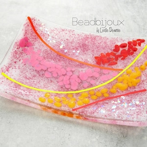 May include: A rectangular, clear glass dish with a pink and white speckled background. The dish features pink, orange, yellow, and red abstract designs. The text "Beadbijoux by Leslie Dearne" is printed on the top right corner.