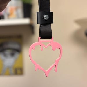 Könnte beinhalten: Ein rosa herzförmiger Anhänger mit tropfendem Design, befestigt an einem schwarzen Riemen mit Metallverschluss. Der Anhänger wird von einem schwarzen Riemen gehalten.