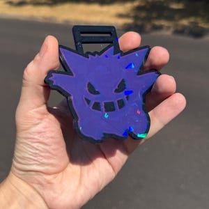 Puede incluir: Una insignia o etiqueta con forma de Gengar morado con un borde negro y un acabado holográfico que refleja la luz. El personaje presenta una amplia sonrisa con dientes y orejas puntiagudas, con un lazo rectangular en la parte superior.