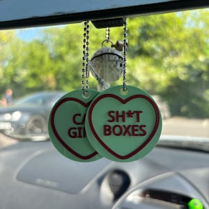 May include: Two green heart-shaped car air fresheners hanging from a rearview mirror. One air freshener says "SH*T BOXES" in red letters. The other air freshener says "C*G*I*N" in red letters.