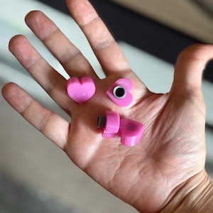Puede incluir: Tres objetos rosas en forma de corazón. Dos son pequeños corazones sólidos, y el tercero es una pieza más grande, en ángulo, con un extremo roscado. Un corazón pequeño tiene un inserto circular negro. Los objetos se sostienen en una mano abierta.