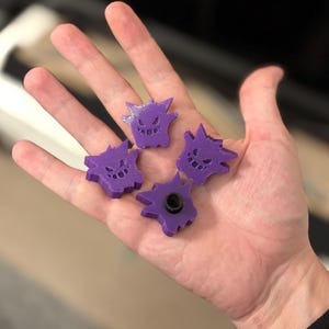 May include: Four purple Gengar figures, a ghost-type Pokémon, are shown. The 3D-printed figures have a cartoon design with jagged edges and smiling faces. One figure has a black screw. The figures are held in a hand.