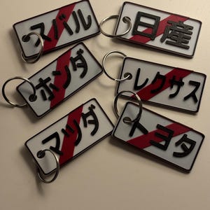 Peut inclure: Six porte-clés blancs avec des rayures rouges et noires. Chaque porte-clés porte un mot japonais différent imprimé dessus : "マビル", "ホシダ", "マリンダ", "ヨダ", "レクサス", et "日露".