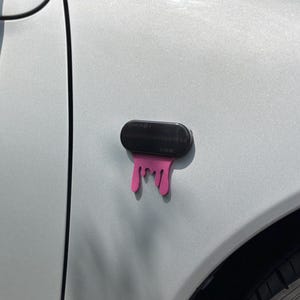 May include: A black car emblem with a pink, dripping design. The emblem is mounted on a light gray car door, near the front tire. The design appears to be a stylized drip.