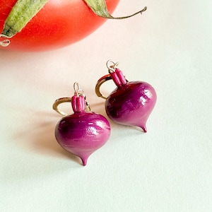 May include: A pair of handcrafted, purple beet earrings with silver-colored ear wires. The earrings are a vibrant shade of purple with a glossy finish, and each has a small, pointed bottom. A red tomato is in the background.