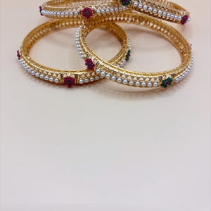 May include: Gold bangles with white pearl accents and colorful gemstone embellishments. The bangles feature red and green gemstones, adding a touch of color to the gold design. These bracelets are a beautiful accessory.