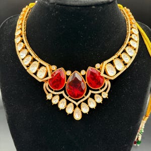 May include: A gold-toned necklace with a central cluster of three large, faceted red gemstones. The necklace features a series of smaller, clear gemstones and is displayed on a black jewelry bust. The necklace has a detailed, ornate design.