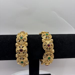 May include: Two gold-toned metal bangles featuring an intricate filigree pattern. Each bangle is decorated with circular motifs containing embedded red and green stones, accented by small clear crystals arranged in a radial design around the center.