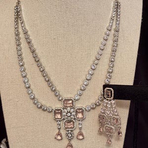 May include: A silver-toned necklace and earring set. The necklace features a double strand design with a central cross-shaped pendant adorned with pink rectangular stones and dangling teardrop-shaped accents. The matching earrings have a similar design.