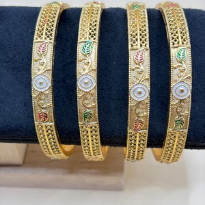 May include: Four gold-colored bangles with intricate filigree patterns. Each bangle features a central white circle with a floral design and is adorned with red and green leaf accents. The bangles are displayed on a black surface.