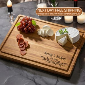 May include: A rectangular bamboo cutting board featuring a grooved edge and a laser-engraved design that reads Avery & Jordan Est. 2025 with a leafy branch motif. The board is topped with a cluster of red grapes, sliced salami, and brie cheese.