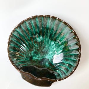May include: A ceramic, scalloped-edge dish with a fan-like pattern in shades of green and brown. The dish has a dark brown rim and a small, curved handle. The glossy finish enhances the colours and texture of the piece.