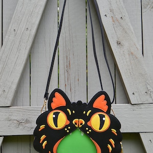 May include: A black crossbody bag shaped like a cat's face with orange and yellow details. The cat's mouth is open and has a green lining.