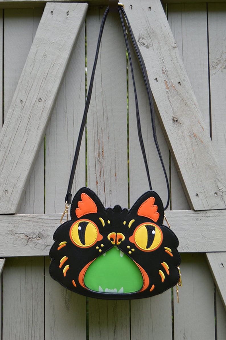 May include: A black crossbody bag shaped like a cat's face with orange and yellow details. The cat's mouth is open and has a green lining.