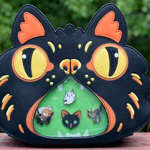 May include: Black cat-shaped purse with a clear plastic window on the front. The window shows a green background with five small pins of various black cats, a ghost, and a pumpkin.