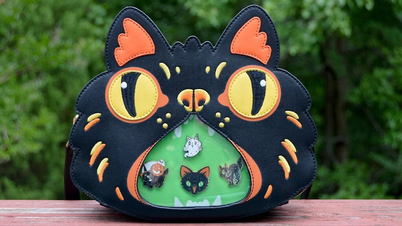 May include: Black cat-shaped purse with a clear plastic window on the front. The window shows a green background with five small pins of various black cats, a ghost, and a pumpkin.