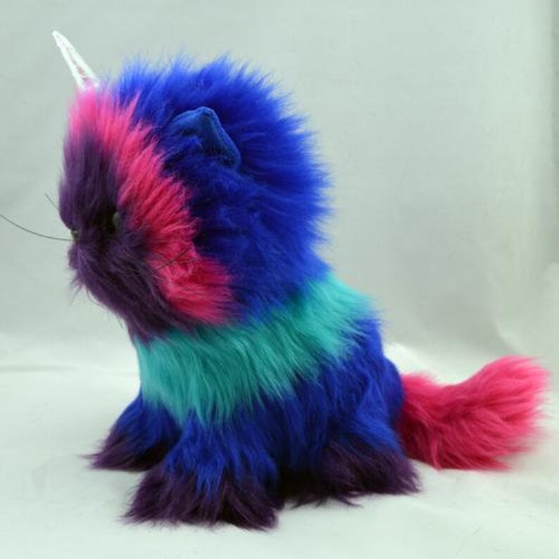 Op de afbeelding: Een pluche kat met een eenhoornhoorn. De kat is bedekt met felgekleurd imitatiebont. Het bont loopt over van paars naar roze, turquoise en blauw. De staart is roze en de hoorn is wit.